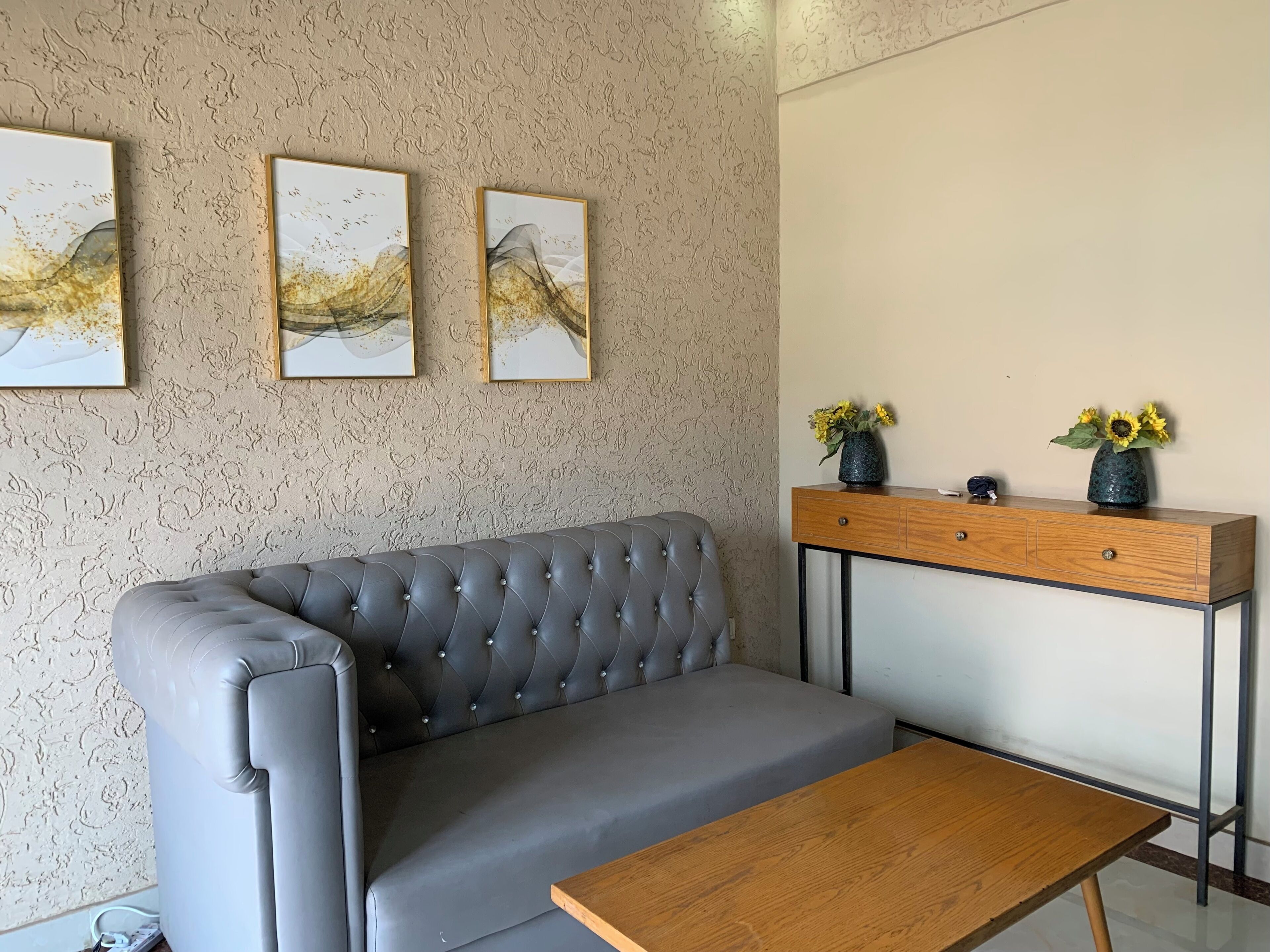 A modern living room with a grey sofa, a wooden coffee table, a wooden side table with two vases of flowers, and three framed artworks on the wall.
