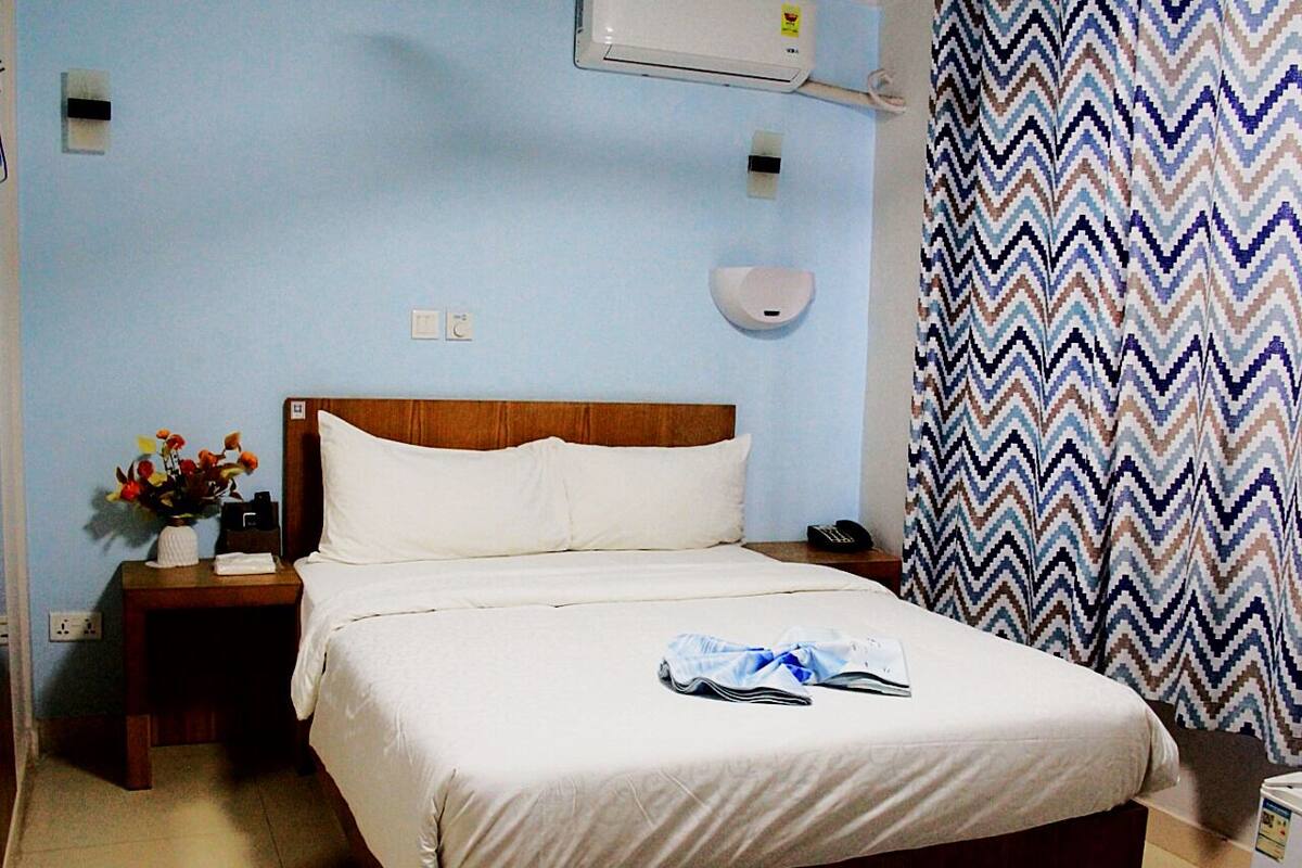 A hotel room with a bed, a nightstand with a vase of flowers, a wall-mounted air conditioner, and a patterned curtain.