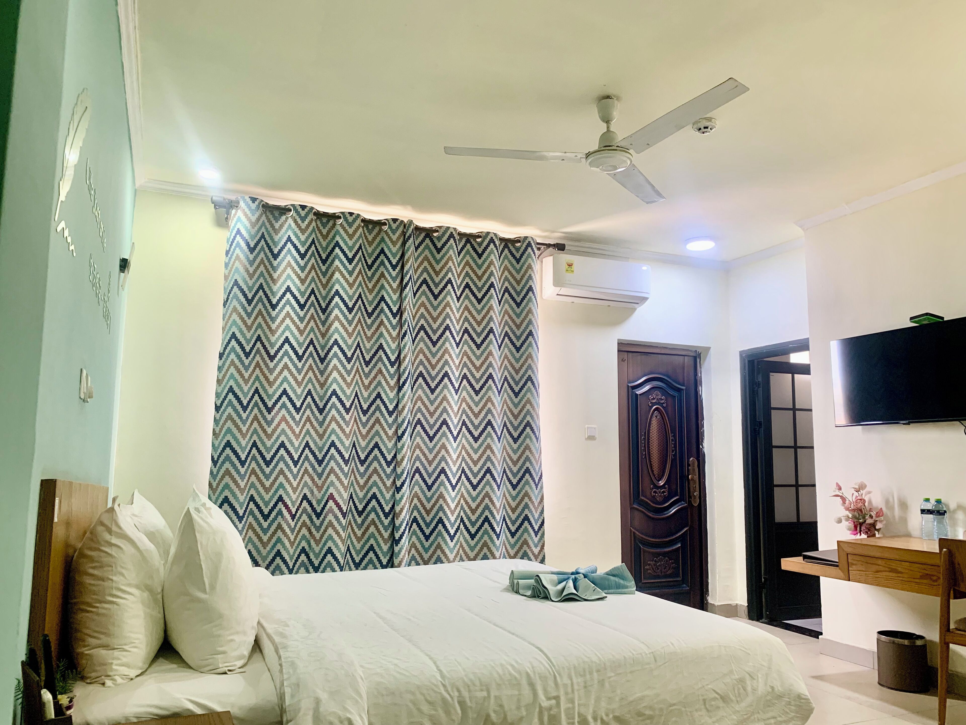 A bedroom with a bed, a ceiling fan, a patterned curtain, a wooden door, and a TV mounted on the wall.