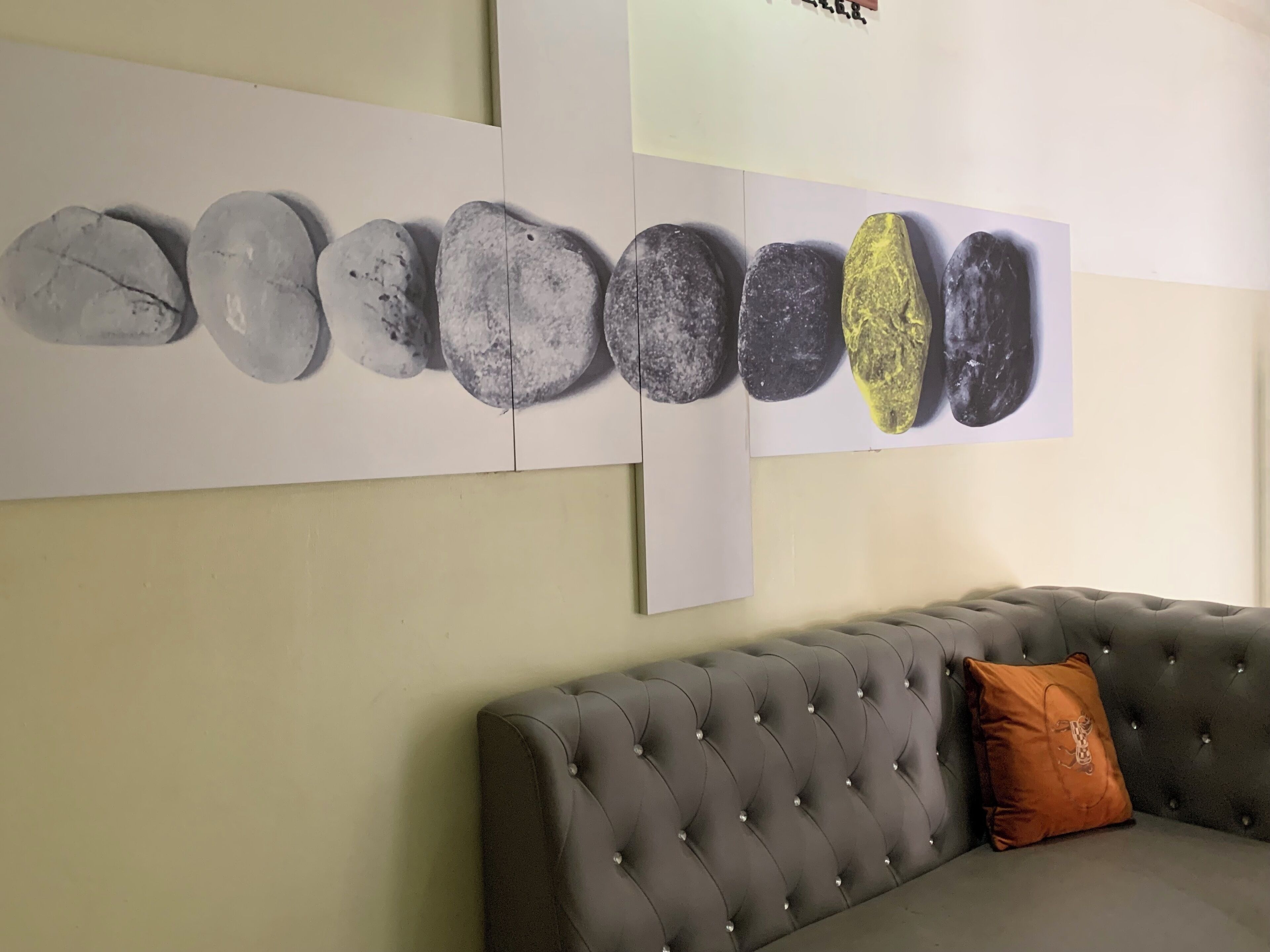 A room with a grey tufted sofa and a cushion, a wall with a series of stone-like images, and a plain wall.