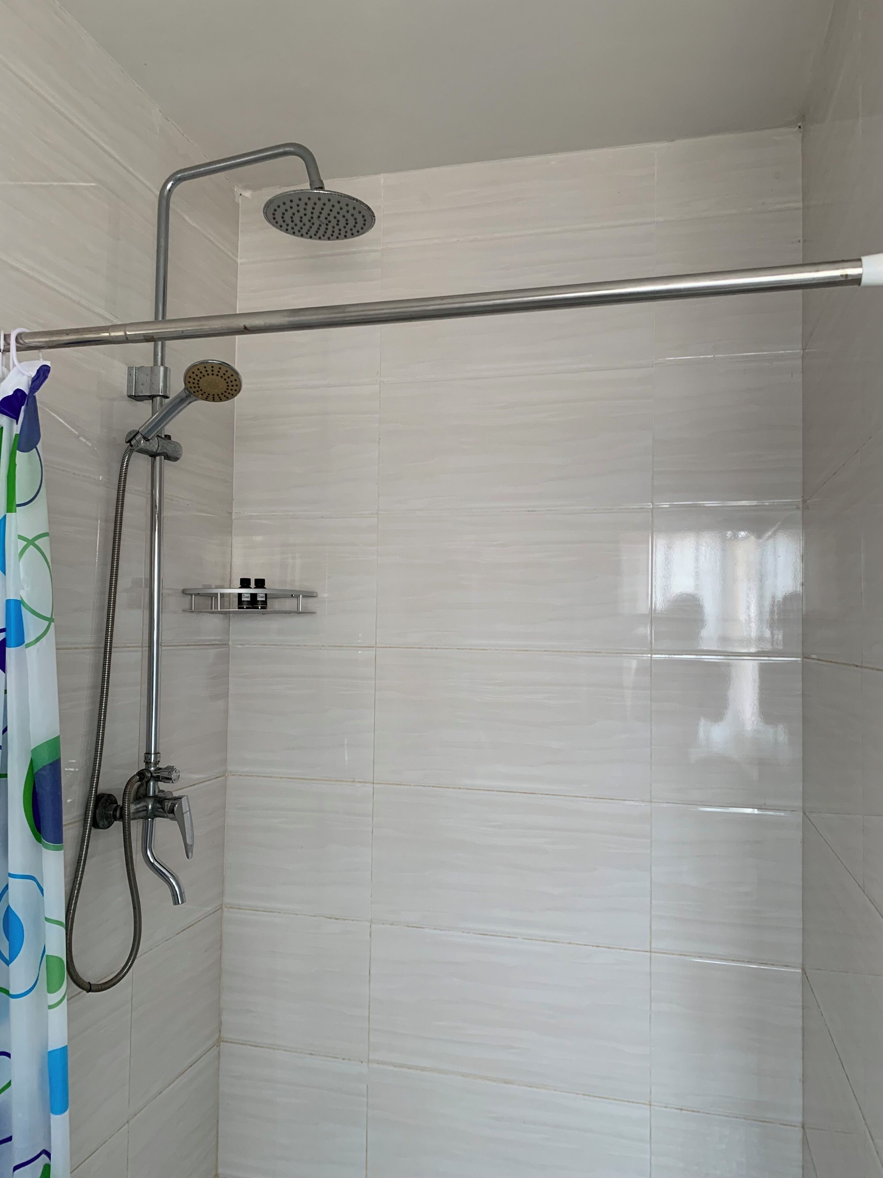 A shower with a handheld showerhead and a fixed showerhead, a metal grab bar, and a small shelf with toiletries.