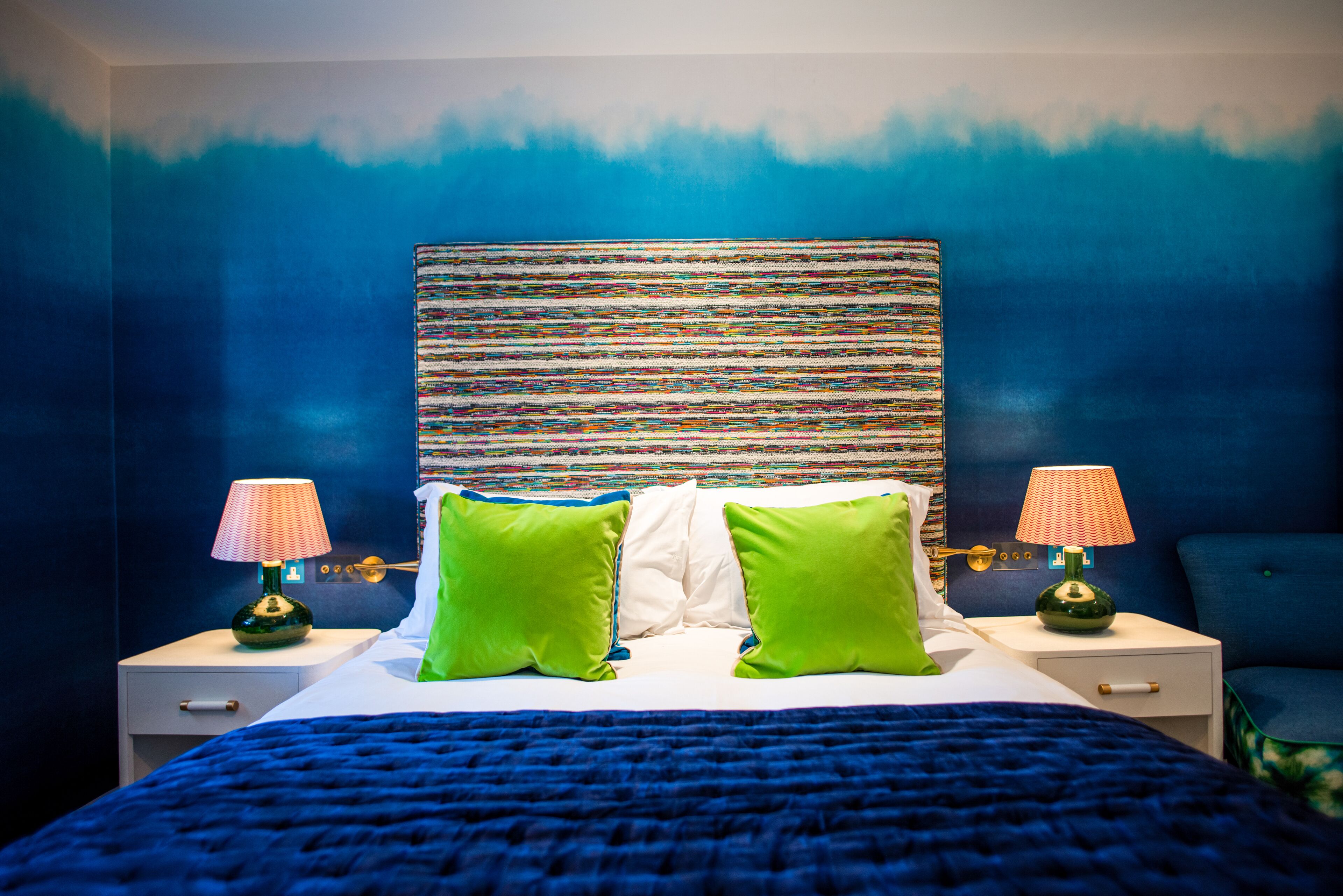 Ein Hotelzimmer mit einem großen Bett, zwei Nachttischen, grün-weißen Kissen und einem farbenfrohen, abstrakten Gemälde an der Wand.