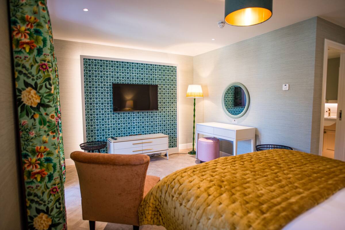 A hotel room with a bed, a desk with a lamp, a TV mounted on a patterned wall, and a chair.