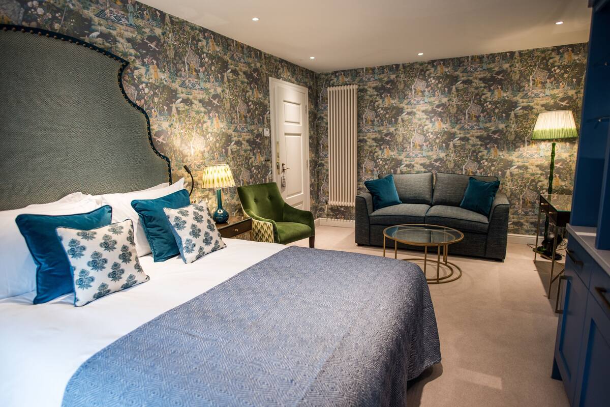 A bedroom with a large bed, a green armchair, a grey sofa, and a small round table.