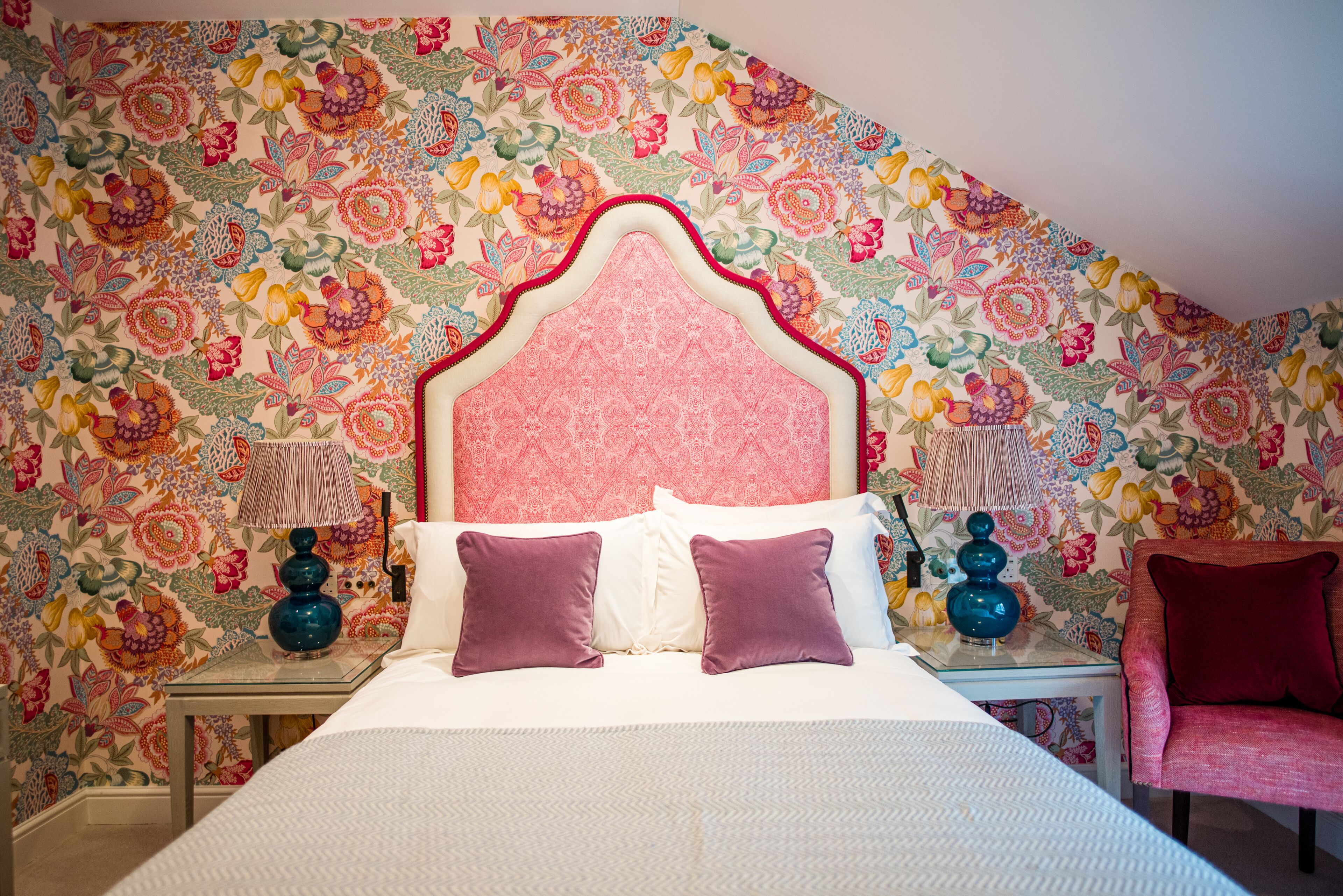 Un dormitorio con papel tapiz floral, una cama con ropa de cama blanca y morada, y una silla roja.