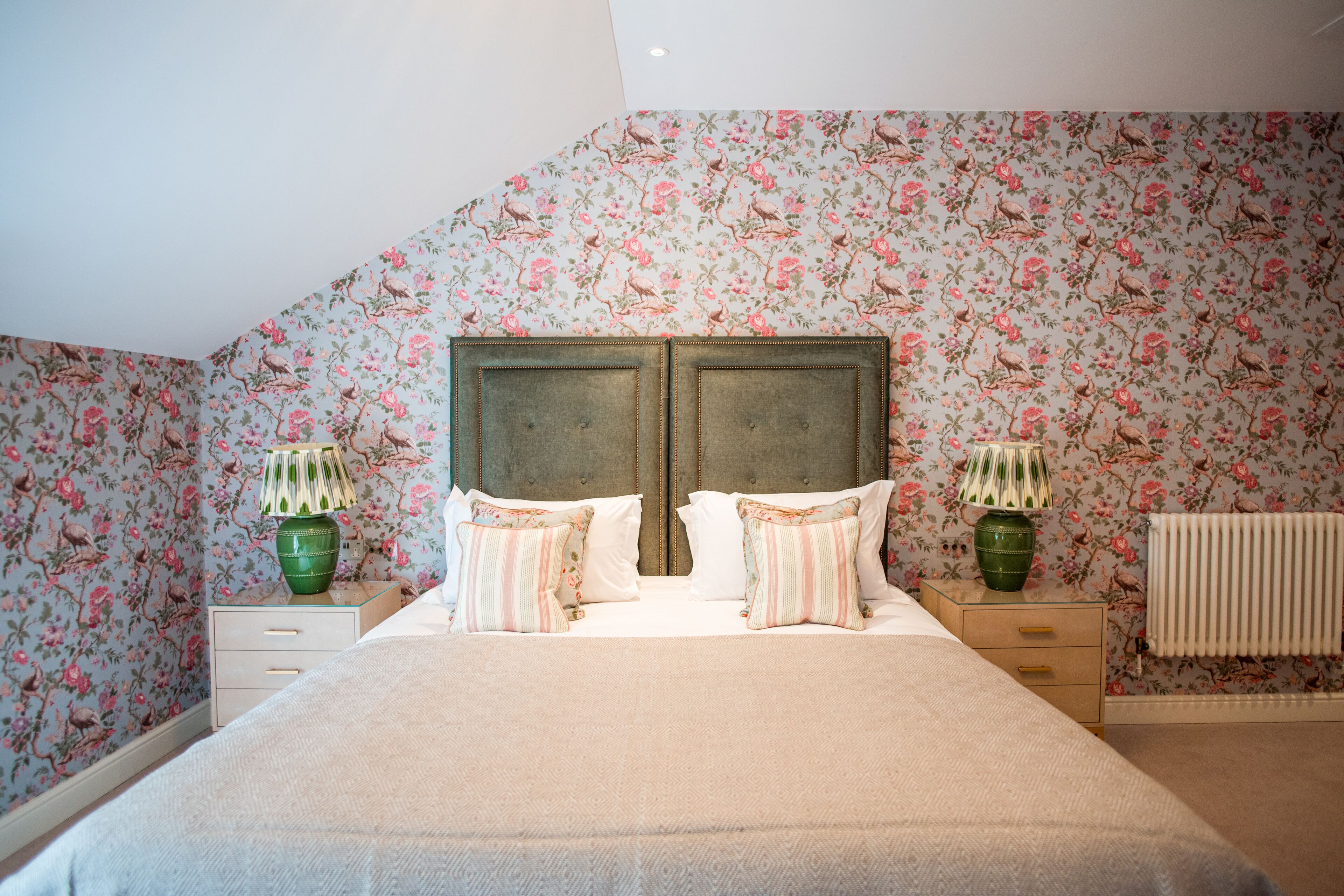 Een slaapkamer met bloemenbehang, een tweepersoonsbed met gestreepte kussens, twee nachtkastjes met lampen en een radiator.