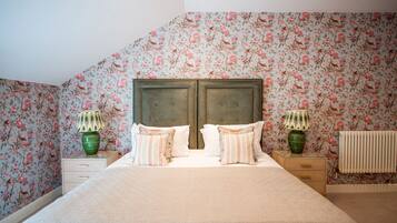 Een slaapkamer met bloemenbehang, een tweepersoonsbed met gestreepte kussens, twee nachtkastjes met lampen en een radiator.
