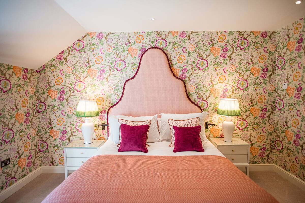 A bedroom with a floral wallpaper, a pink headboard, two bedside tables with lamps, and a bed with a pink blanket and two pillows.