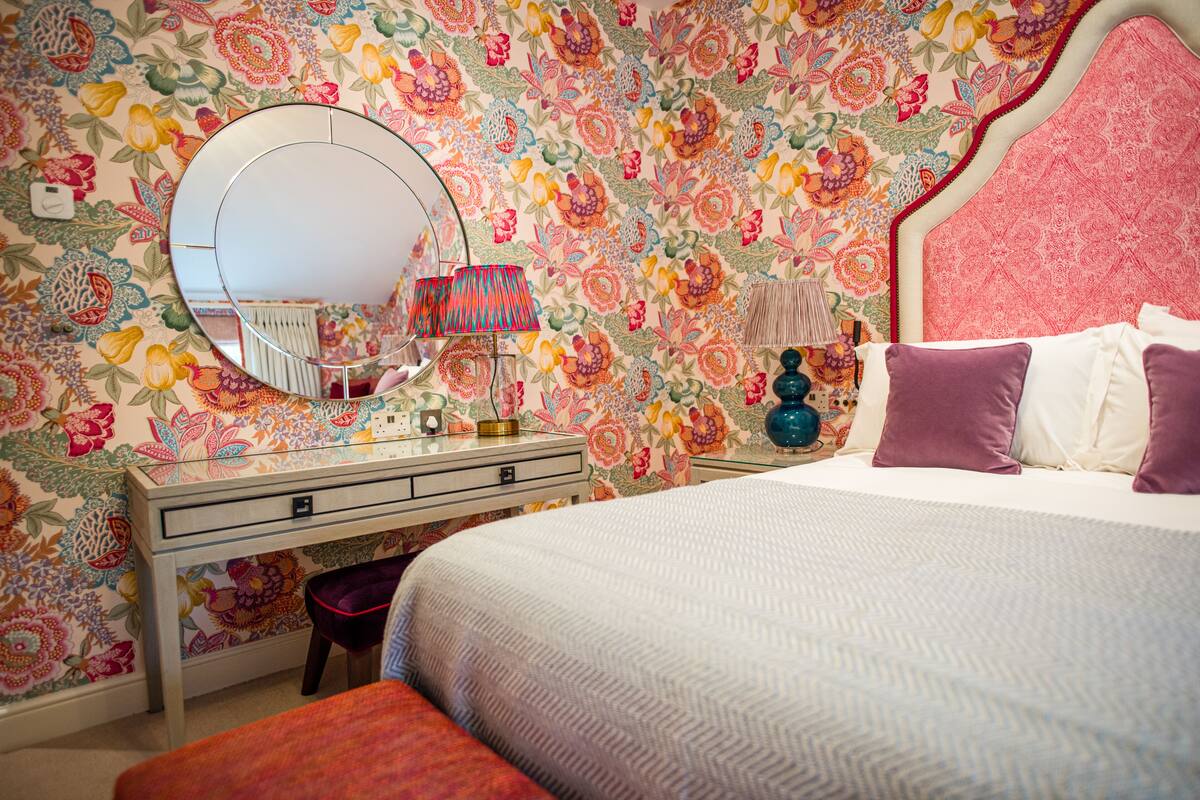A bedroom with a floral wallpaper, a round mirror, a bedside table with a lamp, and a bed with purple pillows.