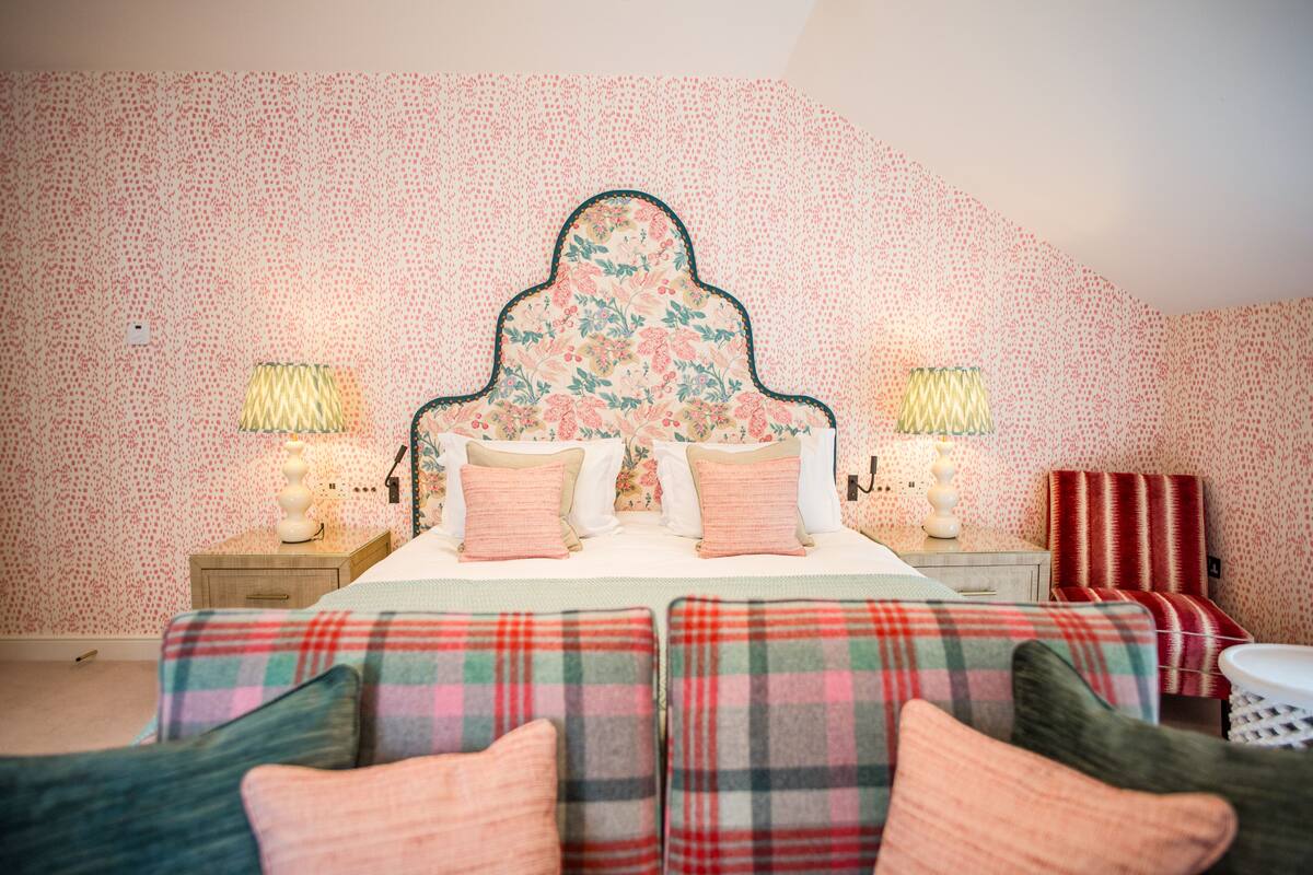 A bedroom with a floral headboard, two bedside lamps, a plaid bedspread, and a red striped chair.