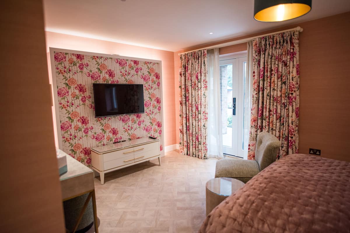 A bedroom with a TV mounted on a floral wallpapered wall, a bed, a chair, and a small table.