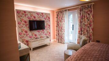 A bedroom with a TV mounted on a floral wallpapered wall, a bed, a chair, and a small table.