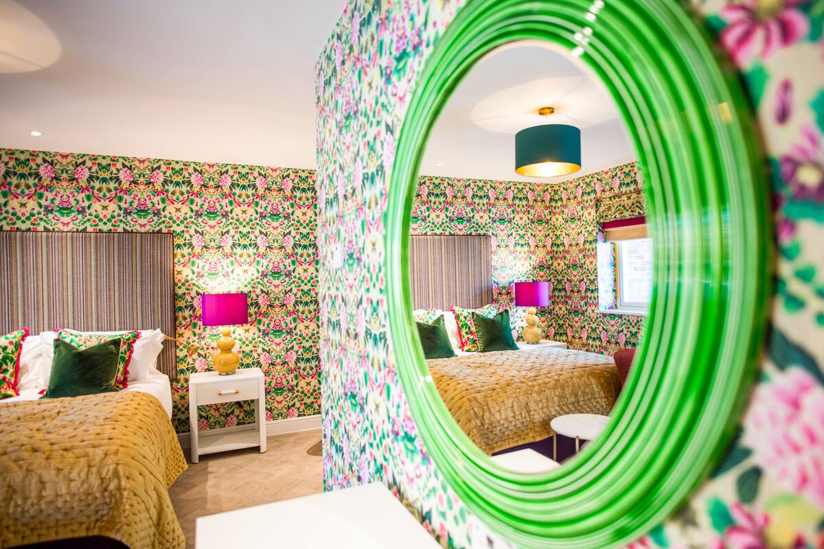 A hotel room with floral wallpaper, a green mirror, two beds, a nightstand, a lamp, and a window.