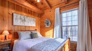 A cozy wooden cabin bedroom with a bed, a nightstand, a wall-mounted artwork, a wreath, and a large window with curtains.