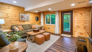 A cozy log cabin interior with a sofa, coffee table, and ottomans, a painting on the wall, and a large window with curtains.