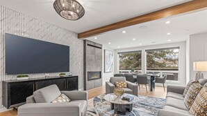 Uma sala de estar moderna com televisão de tela plana, sofás cinzas, uma mesa de centro e um espaço de jantar com cadeiras e mesa.