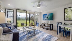 A modern living room with a large sliding glass door leading to a view of a golf course, a ceiling fan, and a dining area with blue chairs.