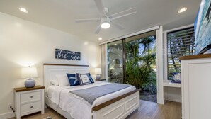 A bedroom with a bed, bedside tables, a ceiling fan, a window with blinds, and a view of greenery outside.