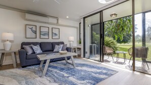 A modern living room with a grey sofa, a coffee table, and a blue rug. There are framed whale paintings on the wall and a large sliding glass door leading to an outdoor patio with chairs.