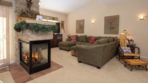 A cozy living room with a stone fireplace, a sectional sofa, a wooden rocking chair, and wall-mounted lights. - Waterside West E206 Fraser 2 bedroom open floor plan & lots of amenities (Fraser)