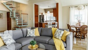 A spacious living room with a gray sectional sofa, a dining area with wooden chairs, a kitchen with a refrigerator, and a staircase with wooden railings.