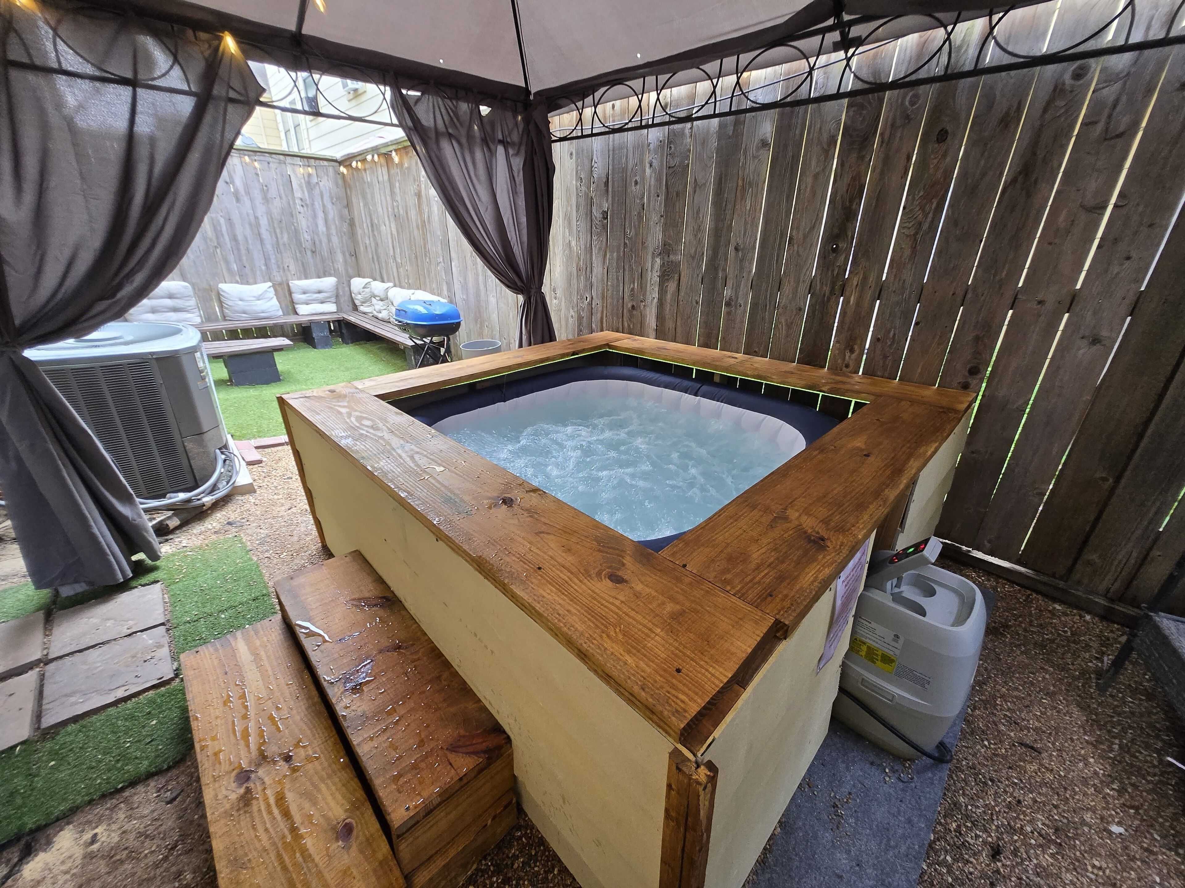 A hot tub with a wooden frame, surrounded by a wooden fence, and a grill in the background.