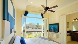 Ein Schlafzimmer mit Bett, Deckenventilator, Fernseher und einem Balkon mit Blick auf Häuser.
