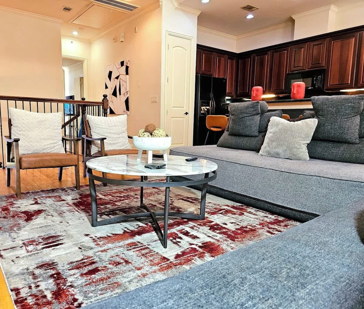A modern living room with a round glass coffee table, a grey sofa with multiple cushions, a wooden chair, and a red and white patterned rug.