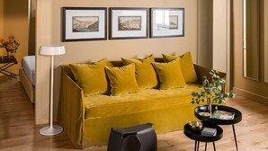 Una sala de estar moderna con un sofá amarillo, una silla negra y una mesita con libros. Hay cuadros en la pared y una lámpara de pie.