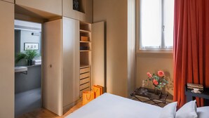Un dormitorio con cama, armario, ventana con cortinas y mesita de noche con un florero.