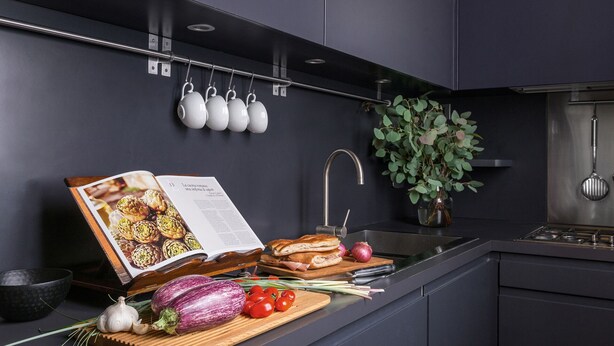 Una cocina moderna con armarios oscuros, un colgador para ollas, una planta en maceta y un libro de cocina abierto sobre la encimera.