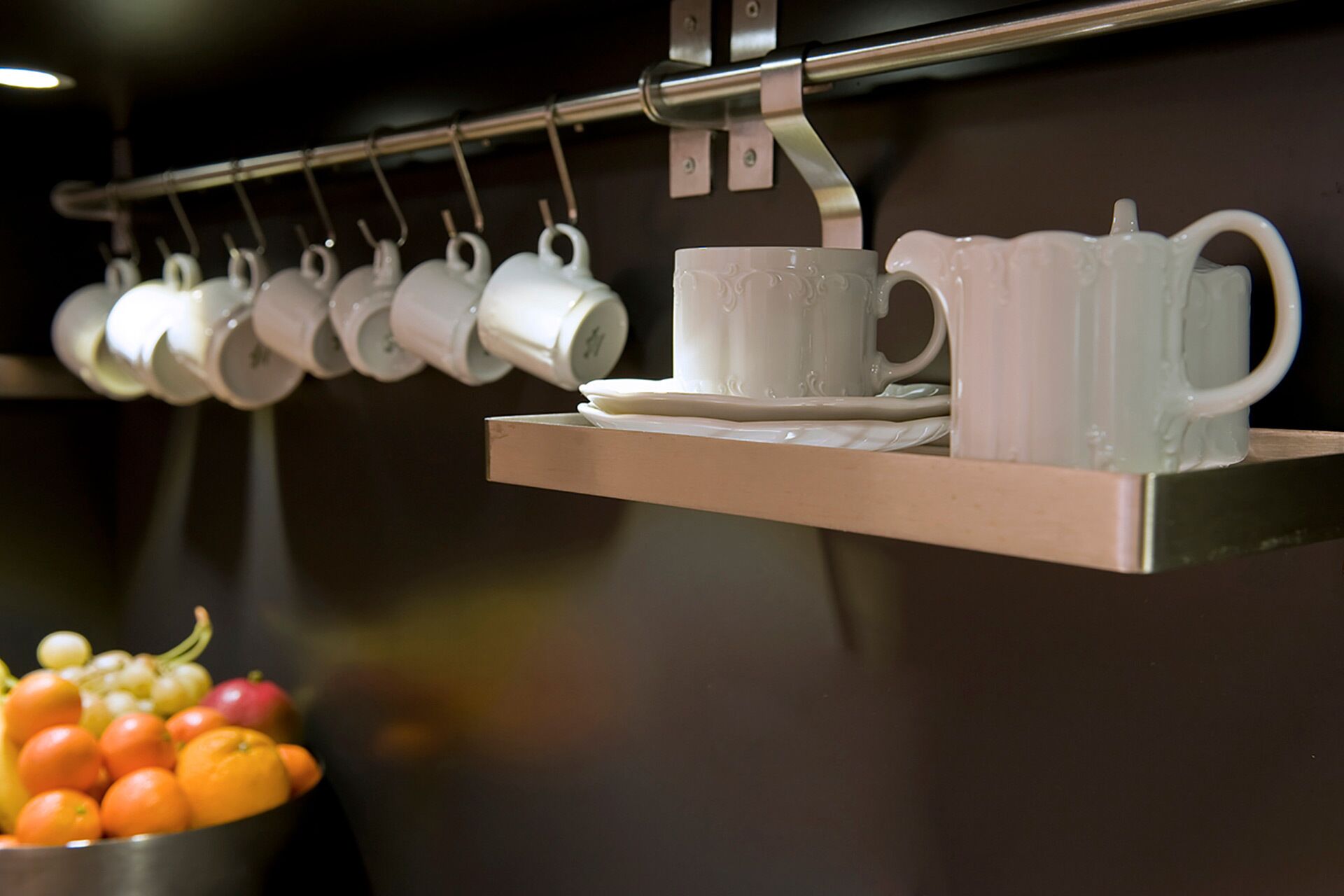 Una cocina con un colgador que sostiene tazas blancas y un frutero sobre una repisa.