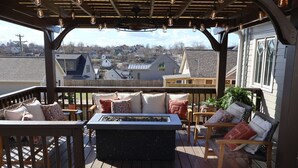 Un espace extérieur aménagé, partiellement abrité, avec un foyer, du mobilier en bois et une vue sur un quartier résidentiel.