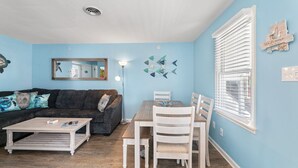 Una sala de estar con sofá, mesa de centro, mesa de comedor y sillas. Hay una ventana con persianas y una decoración en la pared.