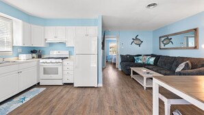 Una cocina compacta con armarios blancos, estufa y refrigerador. En la sala de estar, hay una mesa de comedor de madera y un sofá de color oscuro.