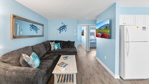 Ein Wohnzimmer mit einem dunkelgrauen Ecksofa, einem Holztisch und einem weißen Kühlschrank.
