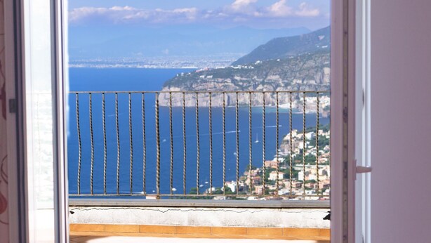 A balcony with a metal railing overlooking a coastal cityscape with a clear blue sea and distant mountains. - Villa Savoia in Sorrento (Sorrento)