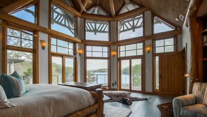 A spacious bedroom with a large bed, wooden headboard, and a view of a lake through large windows. - Parabull's Point, Lake Lure, Luxury House with water slide (Lake Lure)
