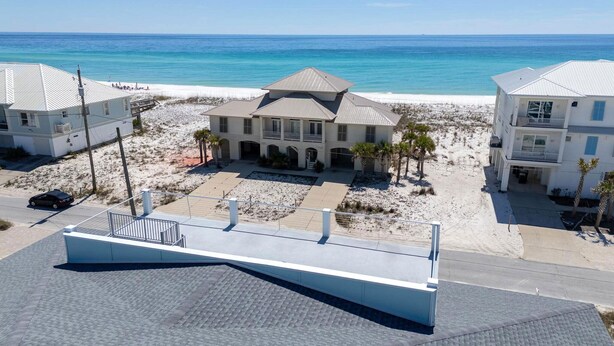 Une propriété située en bord de mer, avec une grande maison dotée d’un balcon, d’une allée et d’une plage de sable en arrière-plan.