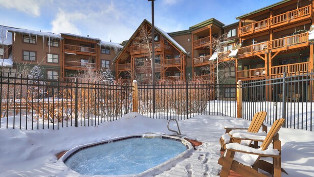 Un espacio exterior cubierto de nieve con un jacuzzi, sillas Adirondack de madera y un edificio de madera con balcones.