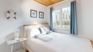 Een slaapkamer met een houten plafond, een bed met witte lakens, een nachtkastje met een lamp en een raam met gordijnen.