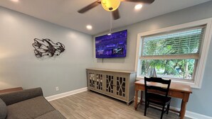 A living room with a gray sofa, a wooden dining table with chairs, a ceiling fan, a mounted TV, and a window with blinds.
