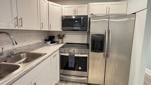 A compact kitchen with white cabinets, a stainless steel refrigerator, a microwave, a stove, and a sink.