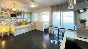 Ein modernes Wohnzimmer mit einer Couch, einem Esstisch mit Barhocker, einem Deckenventilator und einem großen Fenster mit Vorhängen.