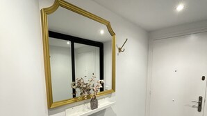 Ein Flur mit einer weißen Tür, einem Spiegel mit goldener Rahmung und einem kleinen Regal mit einer Blumenvase.