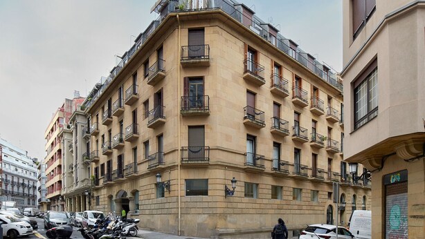 A multi-story building with a corner entrance, balconies, and a street view with parked cars and a person walking. - Marina | The Rentals Collection (San Sebastián/Donostia)