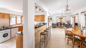 A modern kitchen with wooden cabinets, a central island with stools, a dining area with a wooden table and chairs, and a living room with a sofa and a TV.