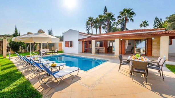 A poolside area with lounge chairs and umbrellas, a dining table with fruit, and a house with a tiled roof and windows.