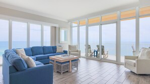 Un salon spacieux avec un grand canapé bleu, une table basse et une vue sur l’océan grâce à de grandes fenêtres.