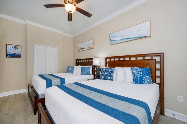 A hotel room with two beds, a ceiling fan, a painting of a beach, and a view of the ocean.